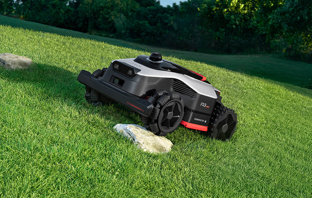 Robot koszący na trawie z widocznymi kamieniami. W tle drzewa i krzewy.