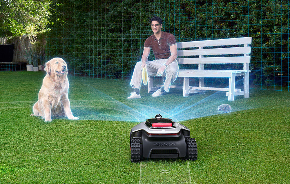 Na trawniku znajduje się robot koszący z widocznymi światłami, a w tle siedzi osoba na ławce obok psa. W tle widoczny jest żywopłot.