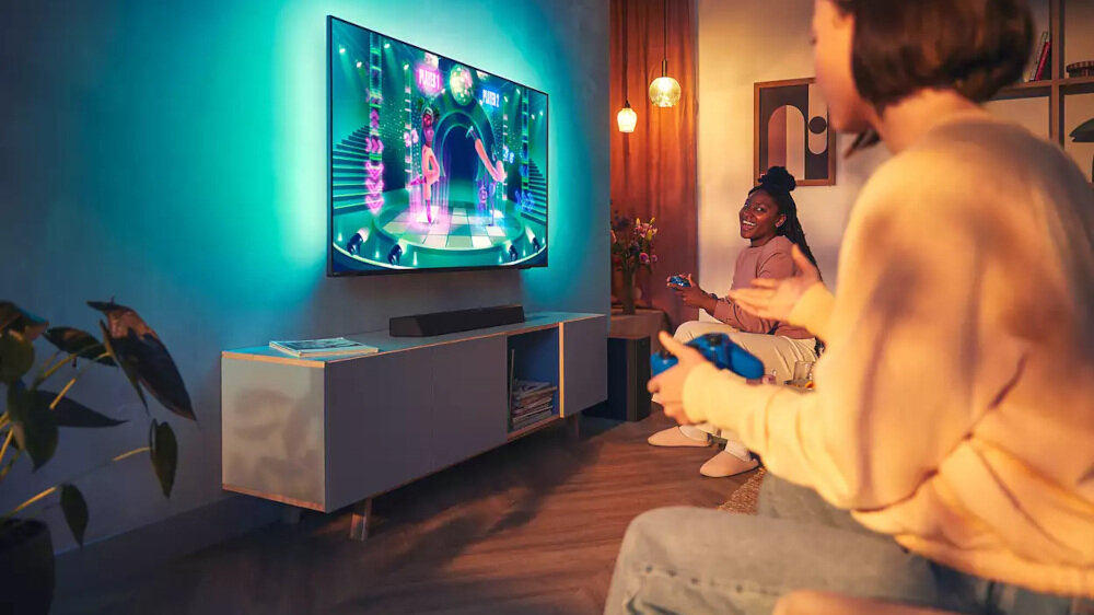 Dwie osoby siedzą w salonie, grając na konsoli przed telewizorem z podświetleniem LED. Na szafce pod telewizorem znajduje się czarny soundbar.