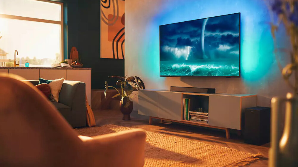 W salonie znajduje się duży telewizor wyświetlający obraz burzy nad oceanem, umieszczony na szafce z książkami i rośliną obok. Pod telewizorem widoczny jest czarny soundbar.