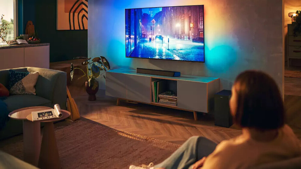 W salonie znajduje się telewizor wyświetlający nocną scenę miejską, umieszczony na szafce z soundbarem poniżej. W pomieszczeniu obecna jest osoba siedząca na sofie, a na stoliku obok leży pilot.