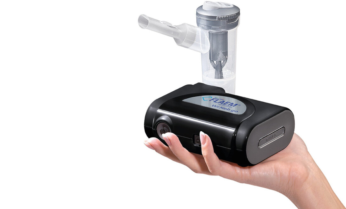 Osoba trzyma czarny inhalator nebulizator z przezroczystym pojemnikiem na górze. Na urządzeniu widoczne jest logo \'FLAEM Wi.Neb Go\'.