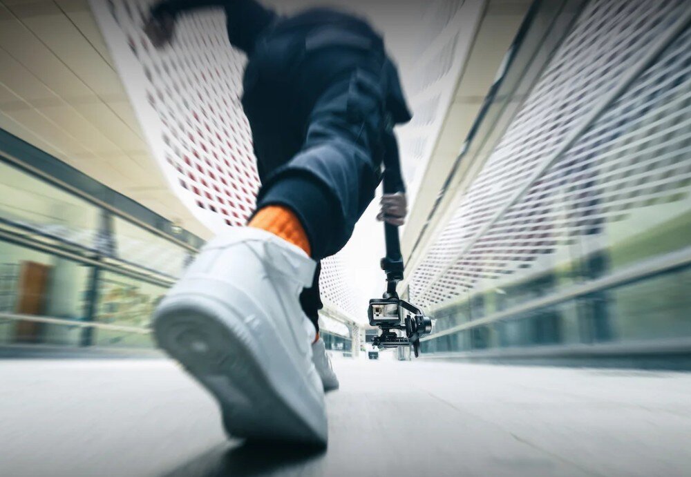 Osoba trzymająca gimbal z kamerą biegnie w nowoczesnym, przeszklonym korytarzu. Widoczne są białe buty i pomarańczowe skarpetki.