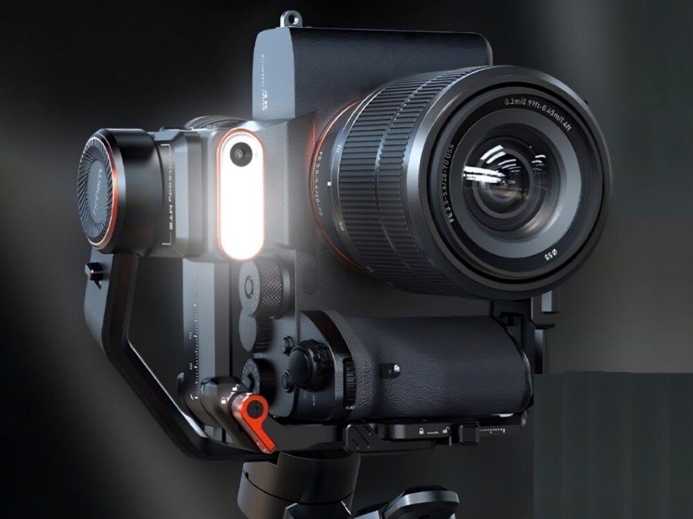 Na obrazie widoczny jest gimbal z zamontowanym aparatem fotograficznym, z dużym obiektywem i podświetleniem LED. Urządzenie ma czarną obudowę z czerwonymi akcentami.