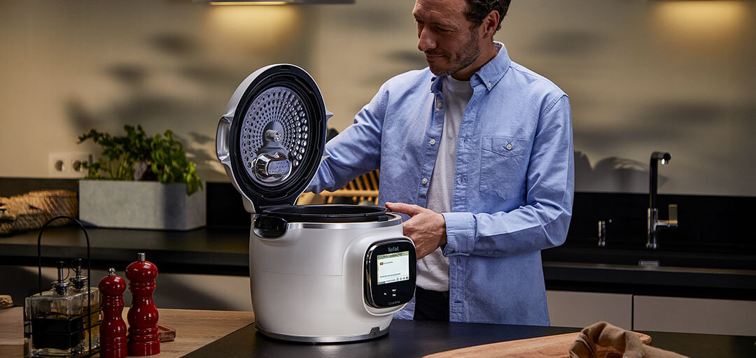 Mężczyzna w niebieskiej koszuli otwiera multicooker marki Tefal w nowoczesnej kuchni. Na blacie obok urządzenia znajdują się czerwone młynki do przypraw i drewniana deska.