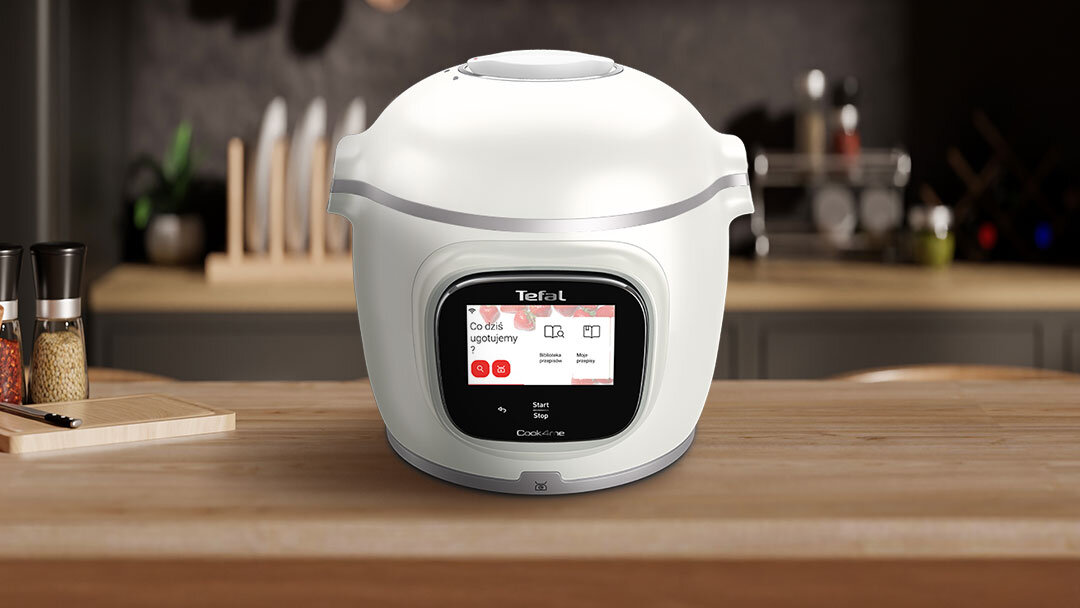 Na blacie kuchennym stoi biały multicooker z wyświetlaczem dotykowym, na którym widoczny jest tekst: \'Co dziś ugotujemy\'. Na urządzeniu znajduje się logo \'Tefal\'.