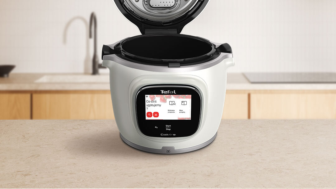 Na blacie kuchennym stoi multicooker z otwartą pokrywą, na wyświetlaczu widoczny jest tekst: \'Co dziś ugotujemy\'.