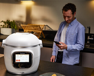 Na blacie kuchennym stoi multicooker z wyświetlaczem, na którym widnieje logo \'Tefal\'. W tle znajduje się osoba trzymająca telefon.