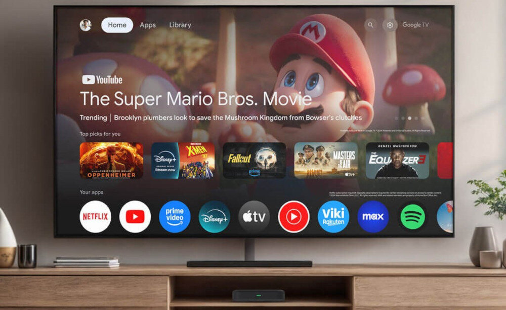 Na ekranie telewizora wyświetlony jest interfejs Google TV z filmem \'The Super Mario Bros. Movie\' oraz ikonami aplikacji takich jak Netflix, YouTube, Prime Video, Disney+, Apple TV, Viki, Max i Spotify. Pod telewizorem znajduje się odtwarzacz multimedialny.