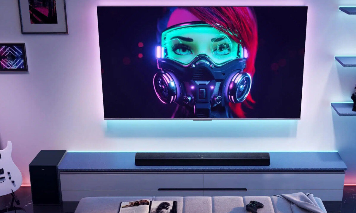 Na ścianie wisi duży telewizor z wyświetlonym obrazem postaci w masce gazowej i goglach. Przed telewizorem znajduje się soundbar na szafce, a obok niego subwoofer.