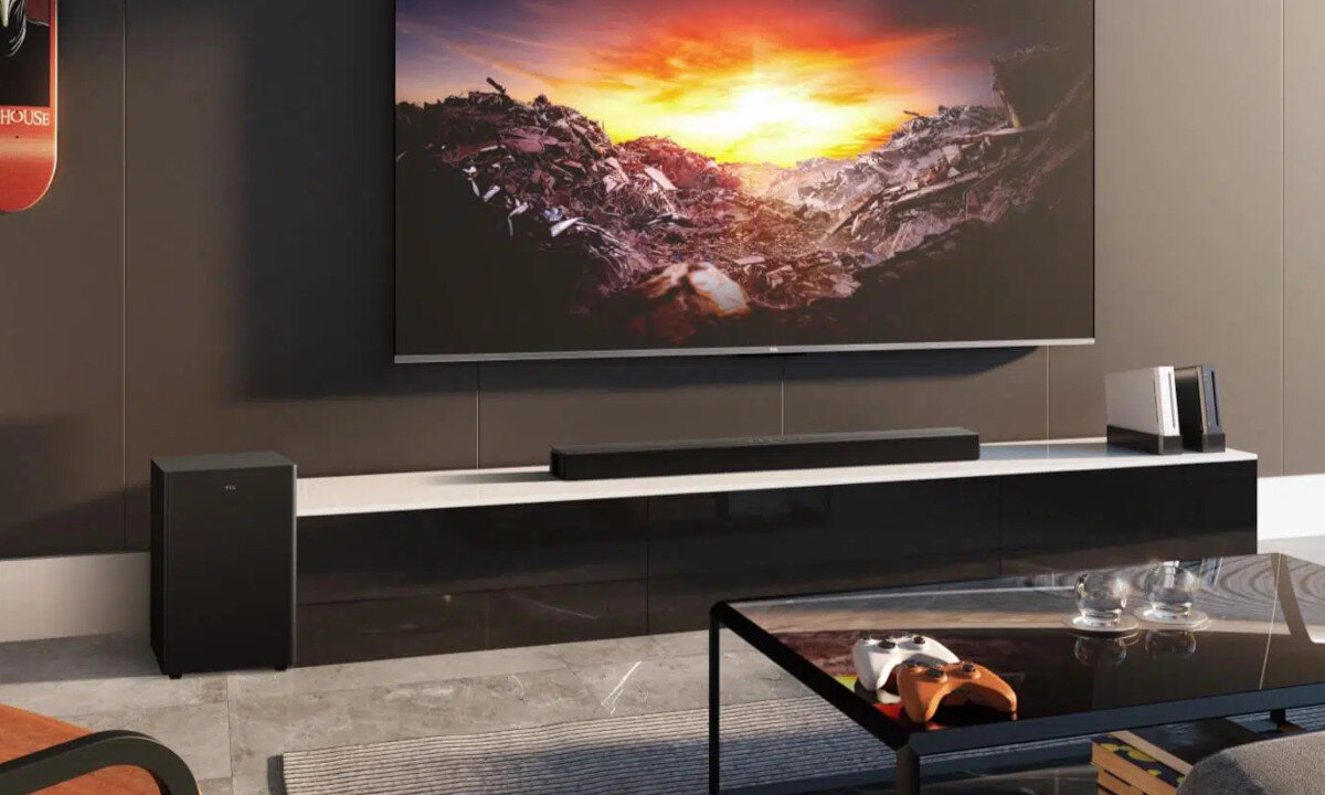 W salonie znajduje się duży telewizor wyświetlający krajobraz z zachodem słońca, umieszczony nad białą szafką, na której stoi soundbar i subwoofer. Na szklanym stoliku przed szafką leży kontroler do gier.