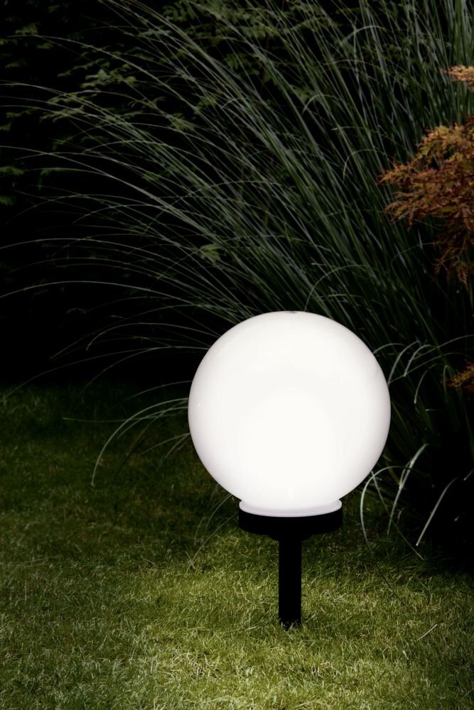 Na obrazie widoczna jest biała, kulista lampa ogrodowa umieszczona na trawniku, otoczona roślinnością. Lampa emituje jasne światło, a w tle widać wysokie trawy i krzewy.