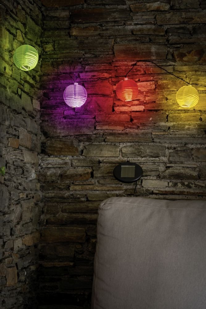 Na tle kamiennej ściany wisi girlanda z kolorowymi lampionami w odcieniach zielonym, fioletowym, czerwonym, pomarańczowym i żółtym. Poniżej znajduje się panel solarny zamocowany na ścianie oraz fragment jasnej poduszki.