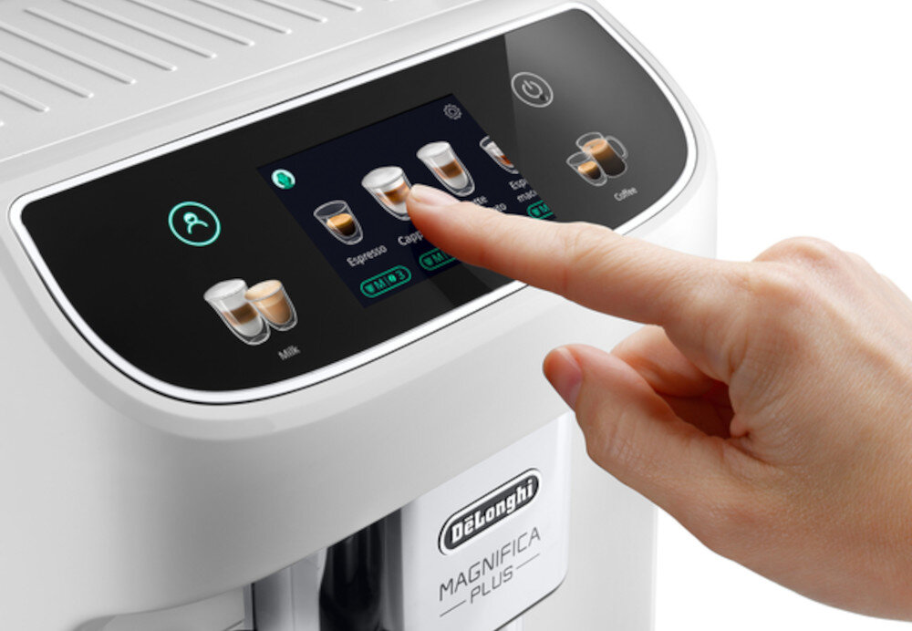 Na zdjęciu widać biały ekspres do kawy z dotykowym panelem sterowania, na którym wyświetlane są różne opcje napojów, takie jak Espresso i Cappuccino. Logo \'DELONGHI MAGNIFICA PLUS\' jest widoczne na przedniej części urządzenia.