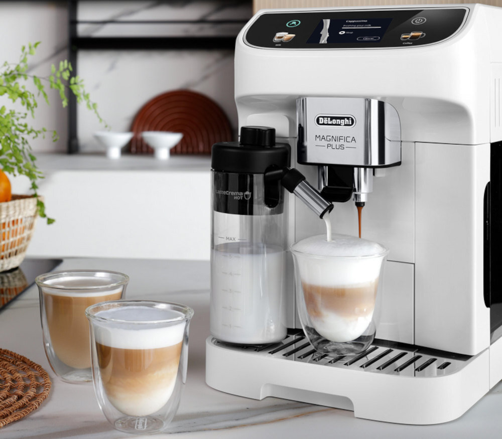 Biały ekspres do kawy z logo \'DeLonghi\' i napisem \'MAGNIFICA PLUS\' przygotowuje kawę do szklanki. Obok stoją dwie szklanki z gotową kawą, a w tle widać elementy kuchenne.