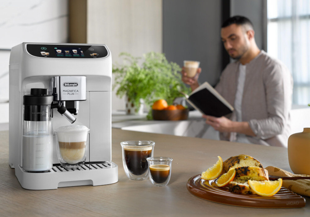 Na blacie kuchennym stoi biały ekspres do kawy z logo \'DeLonghi Magnifica Plus\', obok którego znajdują się trzy filiżanki z różnymi rodzajami kawy. W tle widać osobę trzymającą filiżankę i książkę, a na blacie leży talerz z ciastem i pomarańczami.