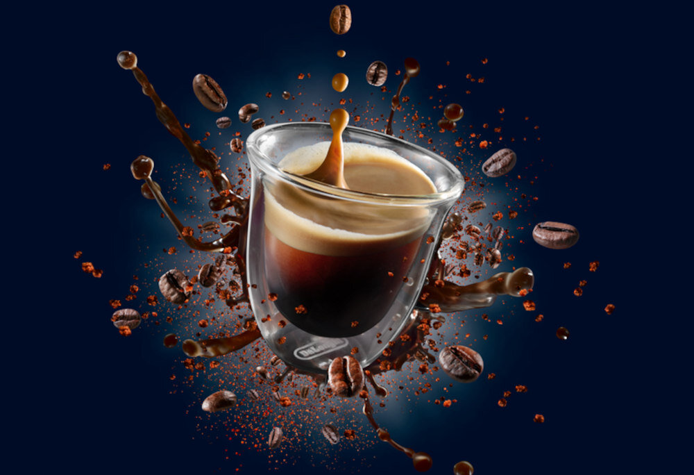 Szklanka z kawą espresso, z której rozpryskują się krople kawy i unoszą się ziarna kawy na ciemnym tle. Logo \'DeLonghi\' widoczne na szklance.