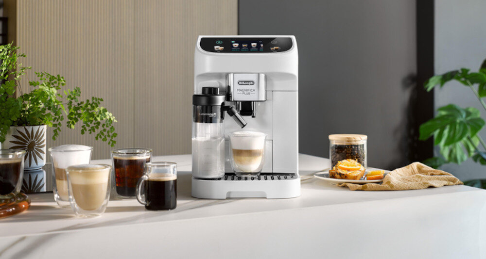 Na blacie stoi biały ekspres do kawy z logo \'DELONGHI Magnifica Plus\'. Obok ekspresu znajduje się kilka filiżanek z różnymi napojami oraz słoik z ciasteczkami.