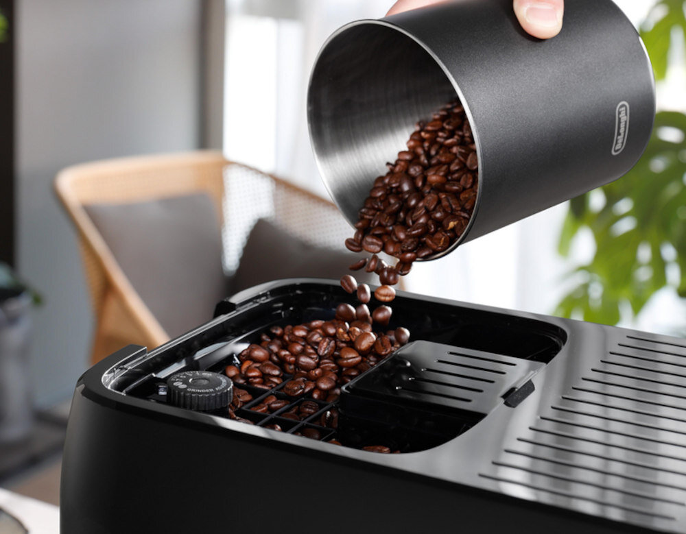 Na zdjęciu widać czarny ekspres do kawy, do którego wsypywane są ziarna kawy z metalowego pojemnika. Na pojemniku widoczne jest logo \'DeLonghi\'.