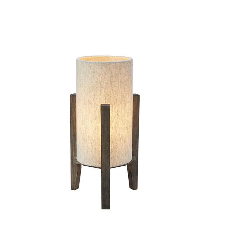 Lampa stołowa o cylindrycznym kloszu w jasnym kolorze, wsparta na trzech drewnianych nogach. Emituje ciepłe, rozproszone światło.