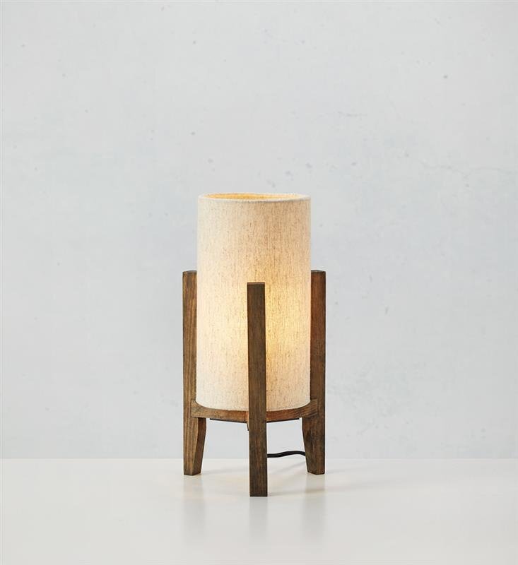 Na obrazie widoczna jest lampa stołowa z cylindrycznym, jasnym abażurem i drewnianą podstawą. Lampa stoi na białej powierzchni, a w tle znajduje się jasna, jednolita ściana.