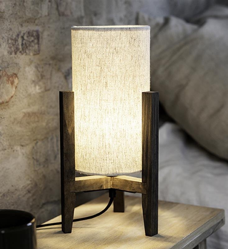 Na zdjęciu znajduje się lampa stołowa z jasnym, cylindrycznym abażurem i ciemną, drewnianą podstawą. Lampa stoi na drewnianym stoliku, a w tle widać ceglaną ścianę i fragment poduszki.