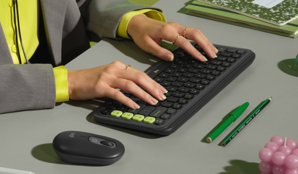 Osoba pisze na czarnej klawiaturze z jasnozielonymi klawiszami funkcyjnymi. Obok znajduje się czarna myszka z logo Logitech oraz zielone długopisy i zeszyty.