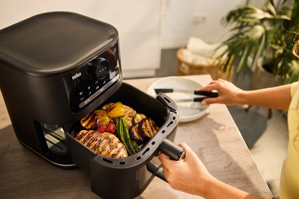 Osoba otwiera kosz frytkownicy beztłuszczowej BRAUN MultiFry, w której znajdują się grillowane warzywa i mięso. Na urządzeniu widoczne jest logo \'BRAUN\'.