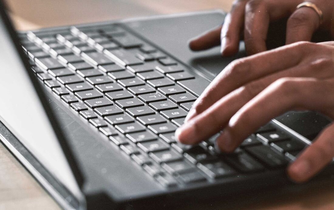 Na zdjęciu widać dłonie piszące na klawiaturze laptopa. Klawiatura ma ciemne klawisze z jasnymi oznaczeniami liter.