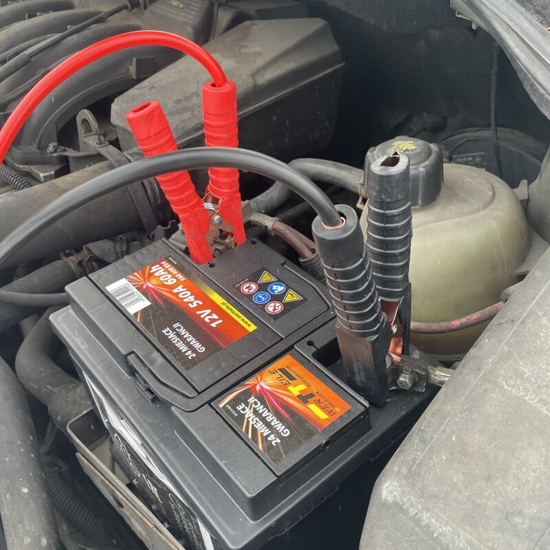Na zdjęciu widać akumulator samochodowy z podłączonymi czerwonymi i czarnymi kablami rozruchowymi. Na akumulatorze widoczny jest tekst: \'12V 540A 60Ah\'.