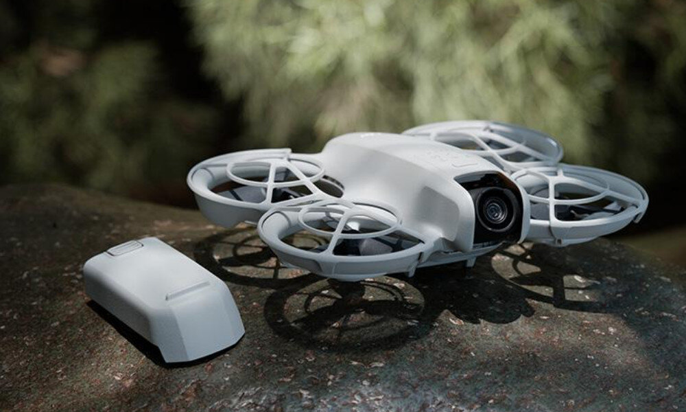 Na obrazie widoczny jest biały dron z kamerą oraz dodatkowa bateria, umieszczone na kamieniu. W tle znajdują się rozmyte elementy roślinności.