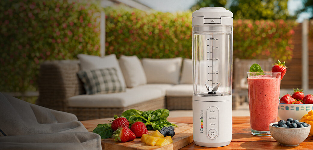 Na drewnianym stole znajduje się biały blender personalny z logo \'icook\', obok którego leży świeże owoce i szklanka z różowym smoothie. W tle widoczna jest kanapa ogrodowa z poduszkami.