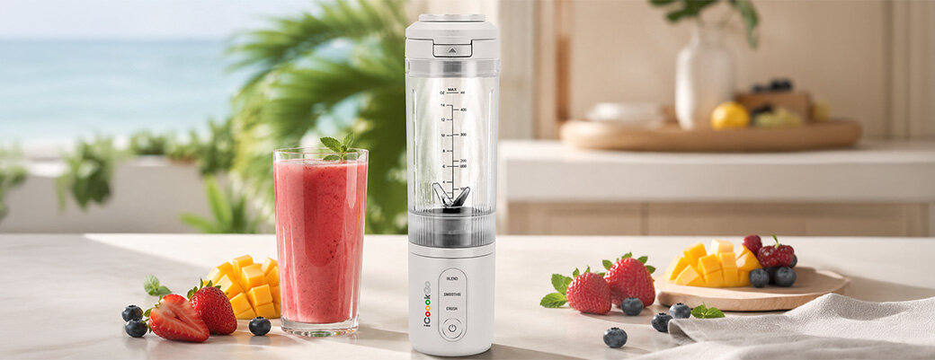 Na blacie kuchennym stoi biały blender personalny z przezroczystym pojemnikiem, obok szklanka z czerwonym smoothie i talerz z owocami. W tle widać rozmyty widok na plażę i morze.