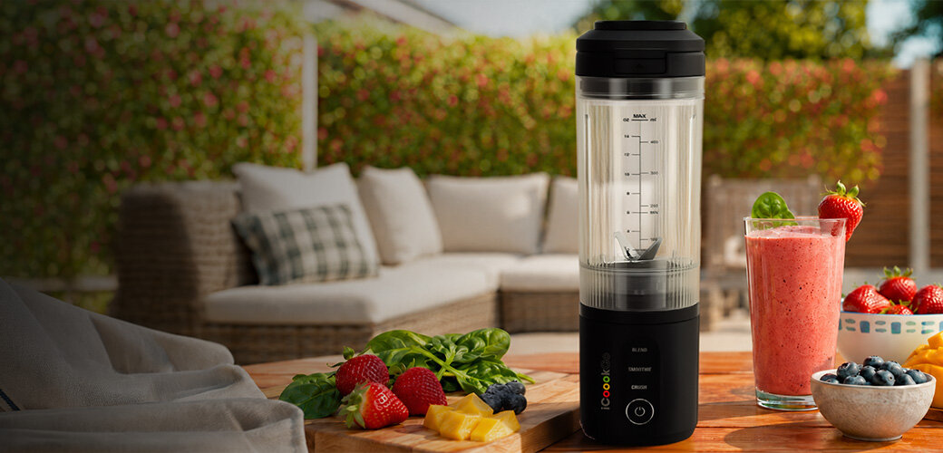 Na drewnianym stole stoi czarny blender personalny ICOOOK z przezroczystym pojemnikiem, obok którego znajduje się szklanka z różowym smoothie oraz świeże owoce i liście szpinaku. W tle widoczny jest ogród z meblami ogrodowymi.