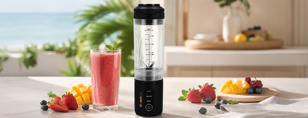 Na blacie stoi czarny blender personalny z logo \'ICOOOK\', obok którego znajduje się szklanka z czerwonym smoothie oraz świeże owoce takie jak truskawki, jagody i mango. W tle widać rozmyty widok na morze i roślinność.