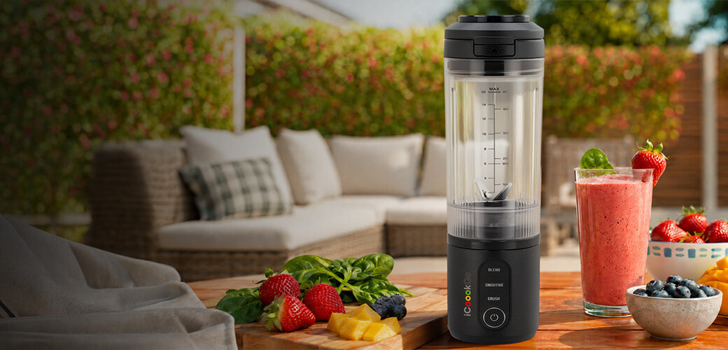 Na drewnianym stole stoi blender personalny w kolorze grafitowym z logo \'icoook\', obok którego znajdują się owoce i szklanka z różowym smoothie. W tle widoczna jest kanapa ogrodowa i zielony żywopłot.