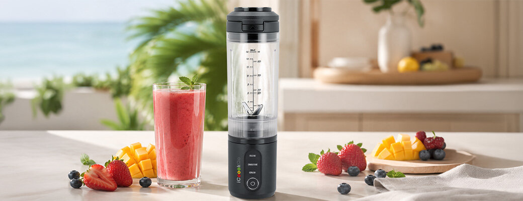 Na blacie kuchennym stoi blender personalny w kolorze grafitowym obok szklanki z czerwonym smoothie. Wokół znajdują się kawałki mango, truskawki, borówki i liście mięty.