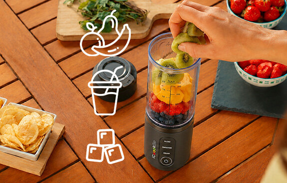Blender iCoook GO MBL-IC-01/B stoi na drewnianym stole, osoba wrzuca do niego owoce. Ikona owoców, kubka i kostek lodu.