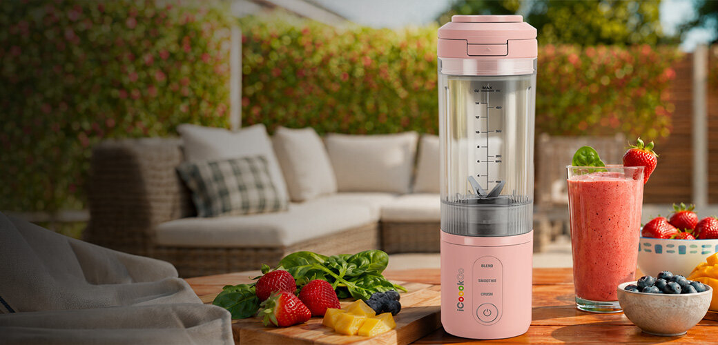 Różowy blender personalny stoi na stole obok szklanki z różowym smoothie i świeżymi owocami. W tle widoczna jest kanapa ogrodowa i zielona roślinność.