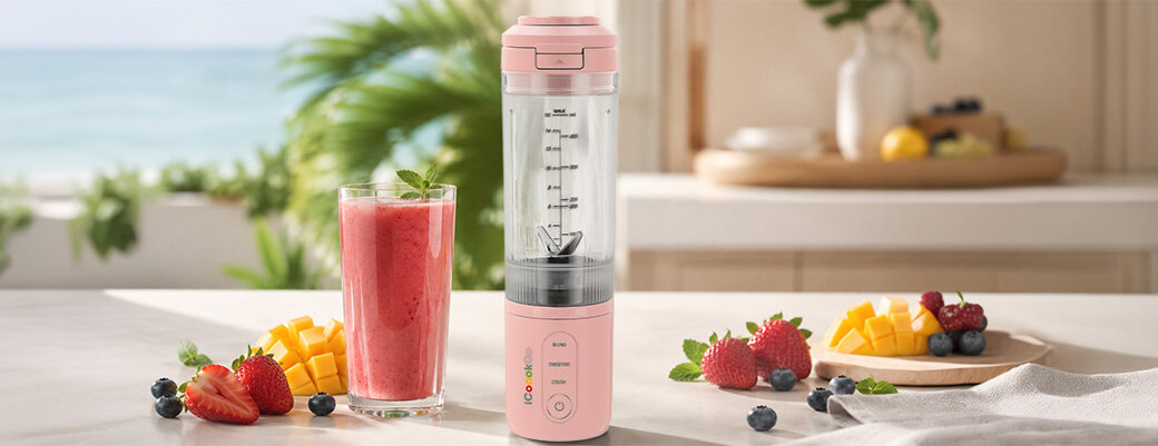 Różowy blender personalny stoi na blacie obok szklanki z czerwonym smoothie i owocami. W tle widać rozmyty widok na plażę i morze.