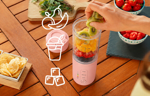 Blender iCoook GO MBL-IC-01/B stoi na drewnianym stole, osoba wrzuca do niego owoce. Ikona owoców, kubka i kostek lodu.
