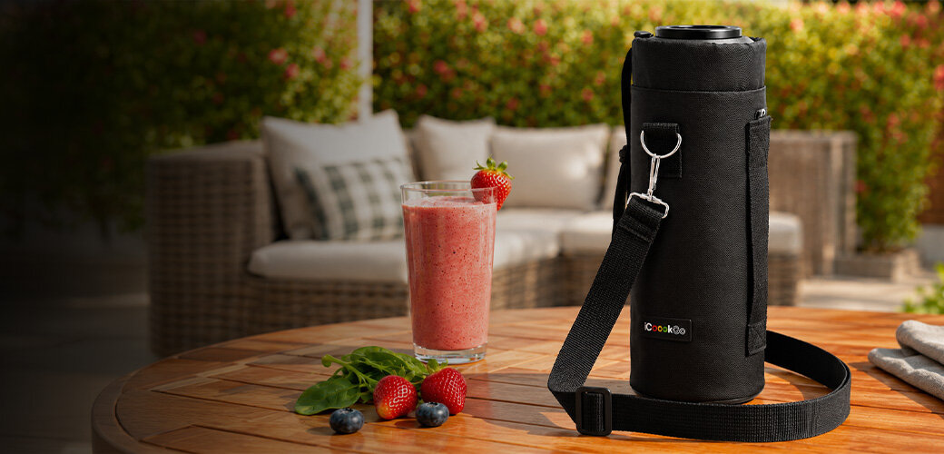 Na drewnianym stole stoi czarne etui na blender z logo \'Cookoo\', obok szklanka z różowym smoothie i kilka truskawek oraz jagód. W tle widoczne są meble ogrodowe i zielone rośliny.