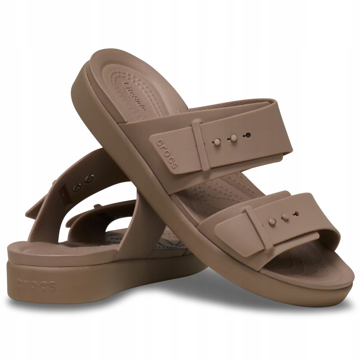 Na obrazie widoczne są beżowe sandały z dwoma paskami i klamrami, z logo \'crocs\' na jednym z pasków. Sandały mają grubą podeszwę i są ustawione pod kątem, jeden na drugim.