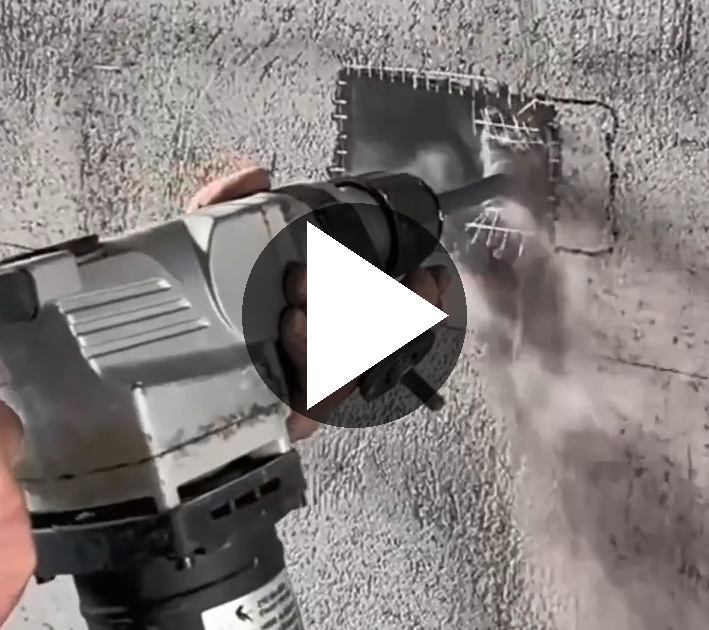 Osoba używa narzędzia elektrycznego do wycinania otworu w betonowej ścianie, generując pył. Na narzędziu widoczny jest duży przycisk odtwarzania wideo.