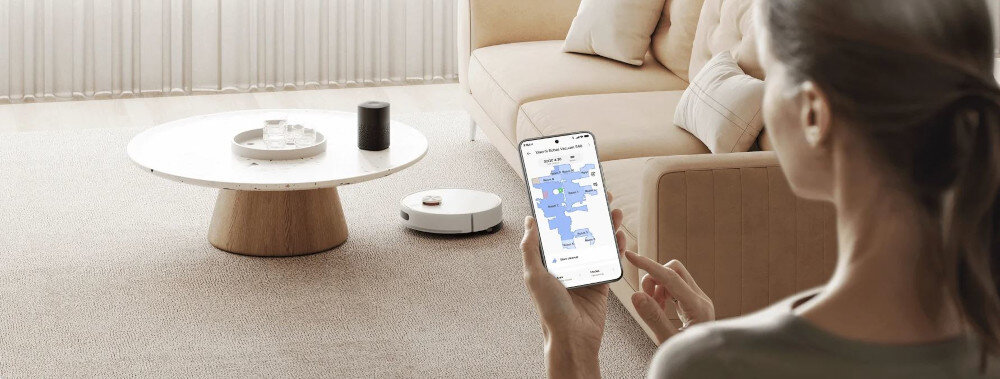 Osoba trzyma smartfon z aplikacją pokazującą mapę pomieszczenia, w tle widać robot sprzątający na dywanie obok stolika z okrągłym blatem.