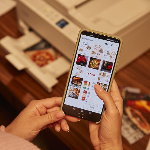 Osoba trzyma smartfon z wyświetlonymi zdjęciami jedzenia, w tle widoczne jest urządzenie wielofunkcyjne z otwartą tacą na papier.