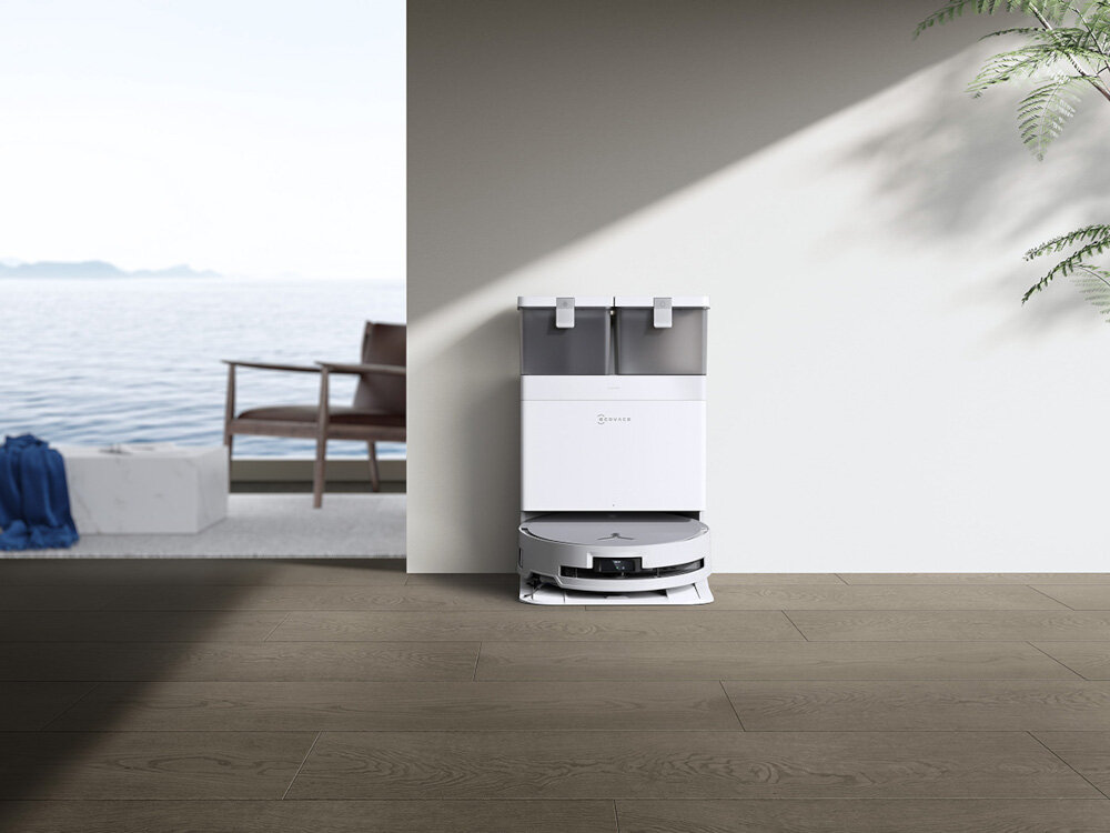 Na obrazie znajduje się biały robot sprzątający ECOVACS Deebot X8 Omni umieszczony przy stacji dokującej na tle jasnej ściany. W tle widać fragment pomieszczenia z widokiem na wodę i meble ogrodowe.