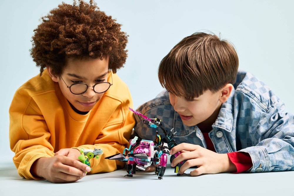 Dwoje dzieci bawi się zestawem LEGO, który przedstawia kolorowego robota z różowymi i czarnymi elementami oraz małą zieloną figurkę. Tło jest jednolite i jasne.