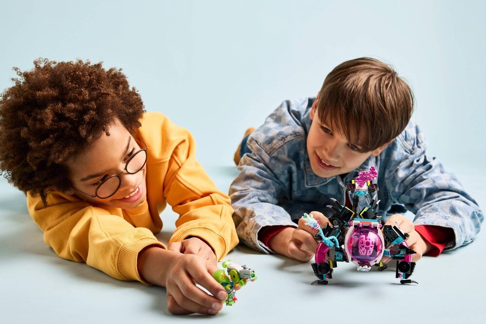 Dwoje dzieci leży na podłodze i bawi się zestawem LEGO, który przedstawia kolorowego robota z różowymi i niebieskimi elementami. Tło jest jednolite, jasnoniebieskie.