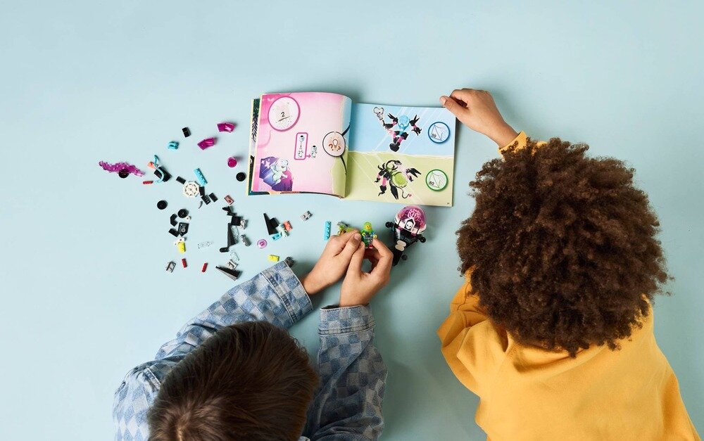 Na jasnoniebieskim tle leżą rozłożone elementy zestawu LEGO oraz otwarta instrukcja z kolorowymi ilustracjami. Dwie osoby składają klocki, trzymając w rękach figurki.